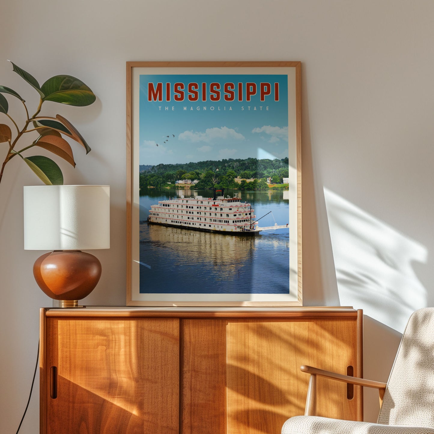 A framed poster of a large white boat on a river, with the text "MISSISSIPPI" and "The Magnolia State" written above it, is displayed on a wooden cabinet in a room with a lamp and a plant.