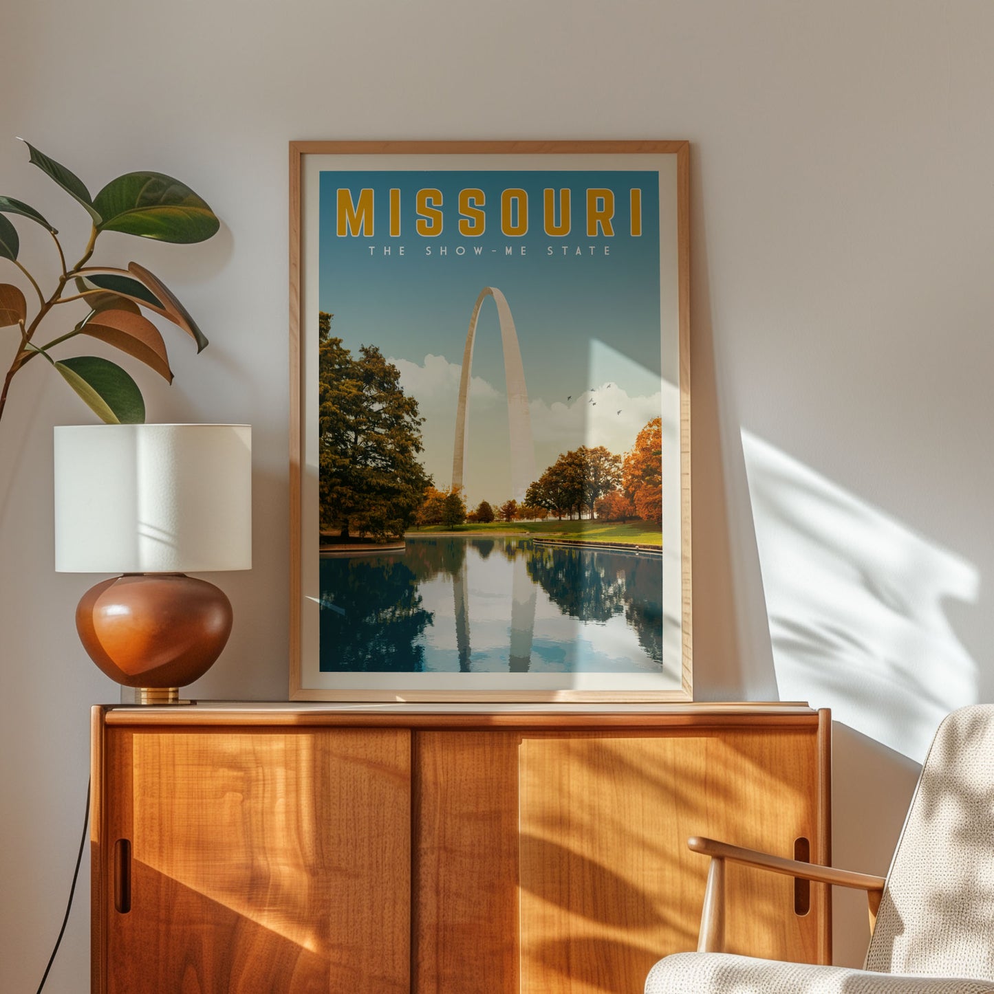 a framed poster of the Missouri arch, which is a large white arch structure located in St. Louis, Missouri. The poster is displayed on a wooden cabinet, and there is a lamp and a plant in the foreground.