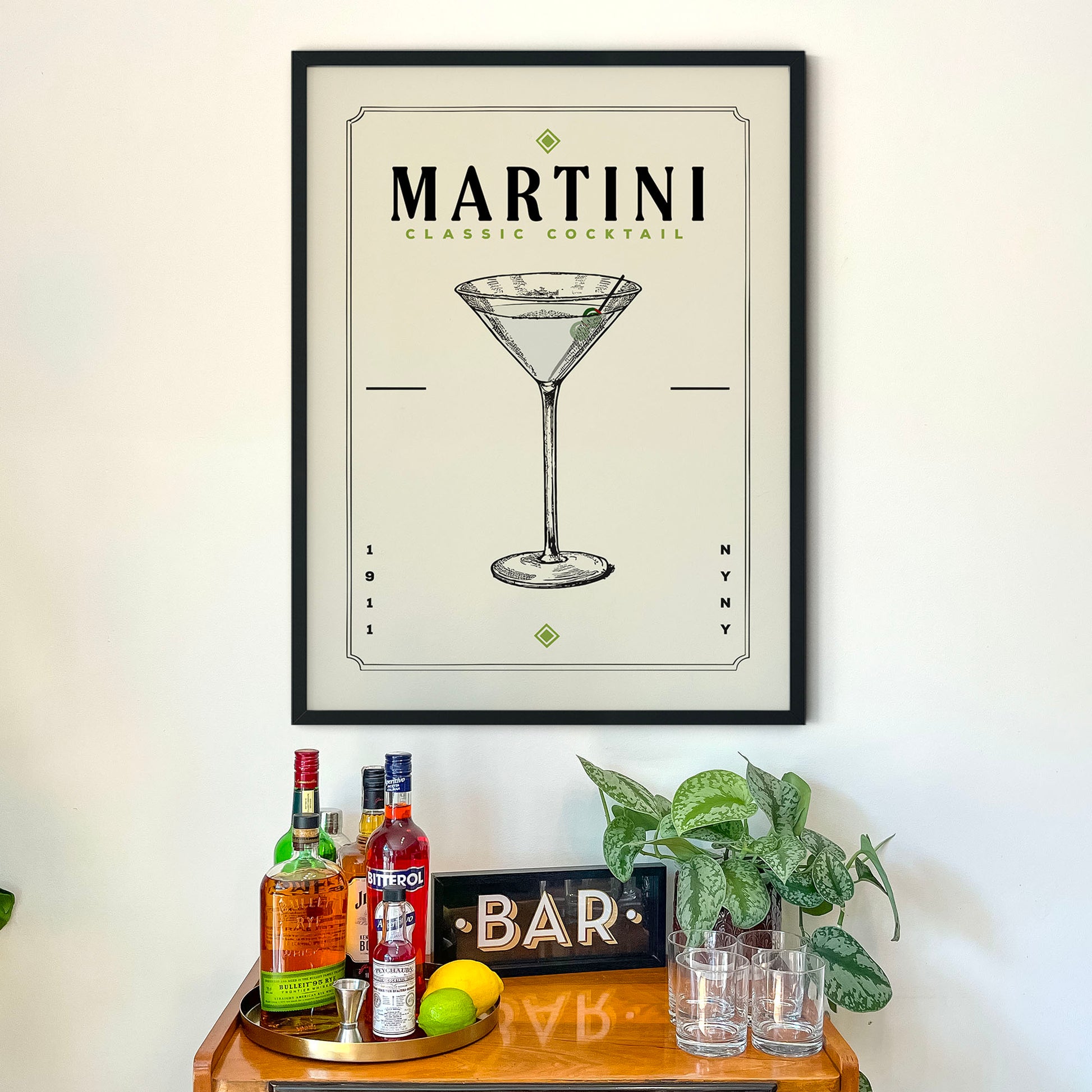 a framed poster of a martini glass on a wall above a wooden bar cart with various liquor bottles and glasses.