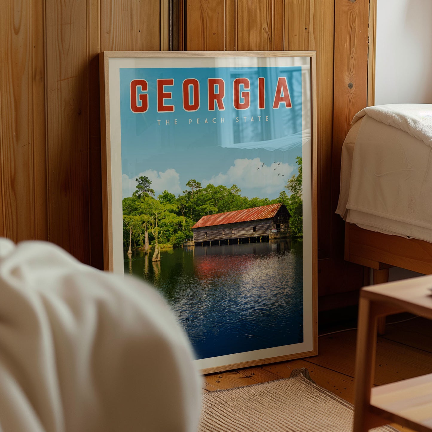 A framed poster of a scenic view of a river with a wooden bridge, surrounded by trees, is displayed in a room with a wooden wall.
