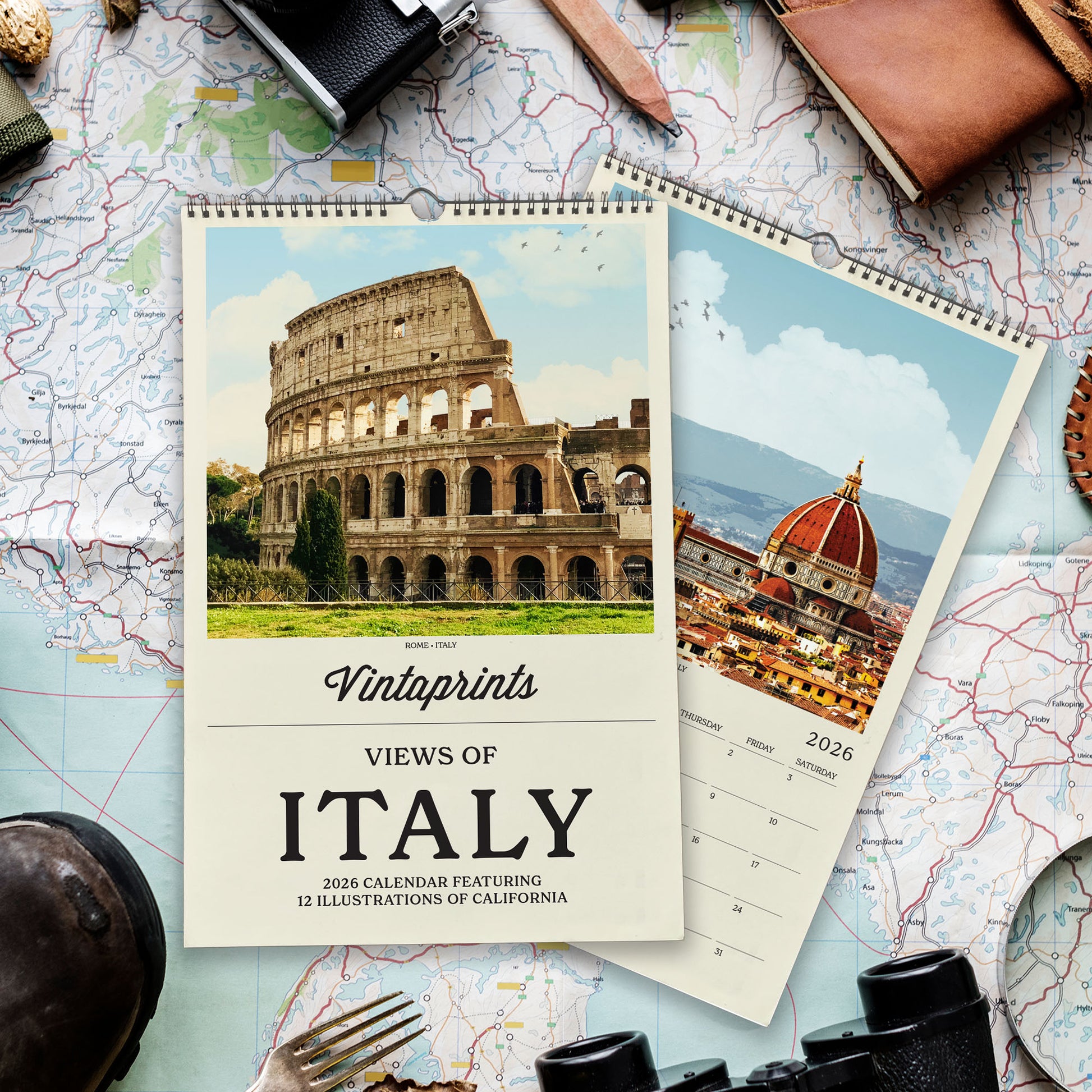 a calendar with a photo of the Colosseum and the Florence Cathedral, along with the text "Vintaprints Views of Italy" and "2026 Calendar Featuring 12 Illustrations of California". The calendar is placed on top of a map, and various travel-related items such as a camera, a hat, and a magnifying glass are also visible in the image.