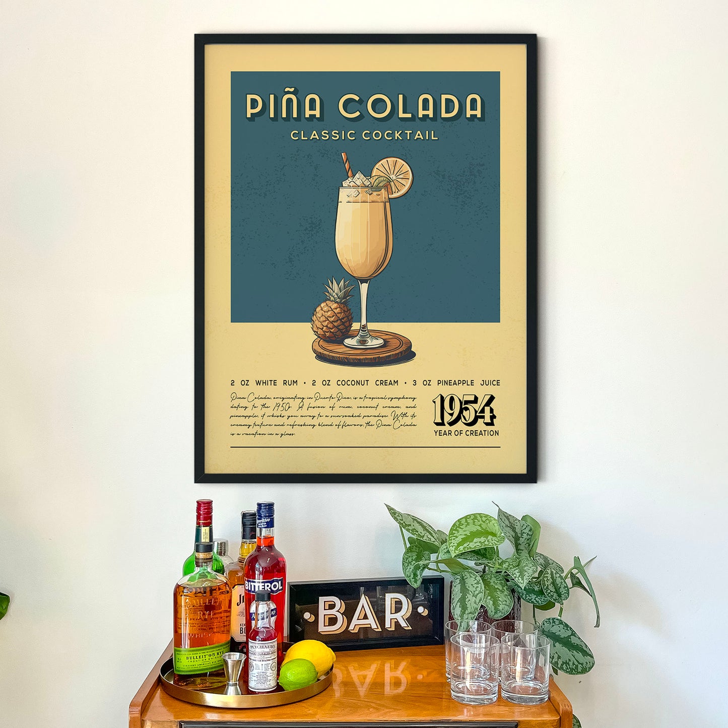 a framed poster of a classic cocktail, specifically a Pina Colada, hanging on a wall above a wooden bar counter. The poster includes illustrations of the cocktail and its ingredients, such as a pineapple, a glass of rum, and a coconut cream. The bar counter has various bottles of alcohol and a plant, creating a cozy and inviting atmosphere.