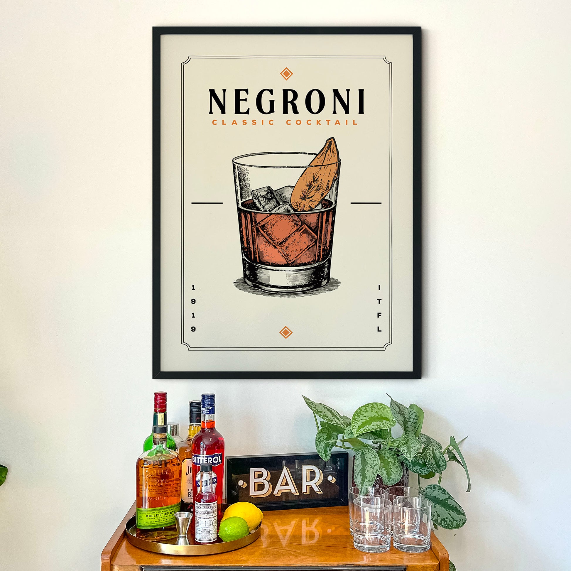 a framed poster of a cocktail glass with a cocktail, hanging on a wall above a bar counter with various liquor bottles and a plant.