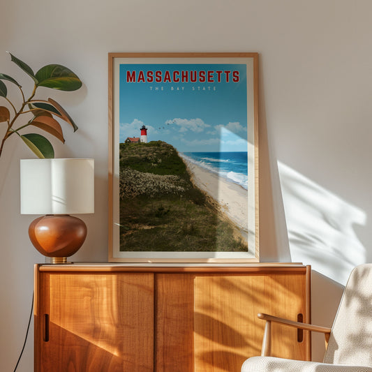 a framed poster of a lighthouse on a beach, with the text "MASSACHUSETTS THE BAY STATE" written above it. The poster is displayed on a wooden cabinet, and there is a lamp and a plant in the foreground.