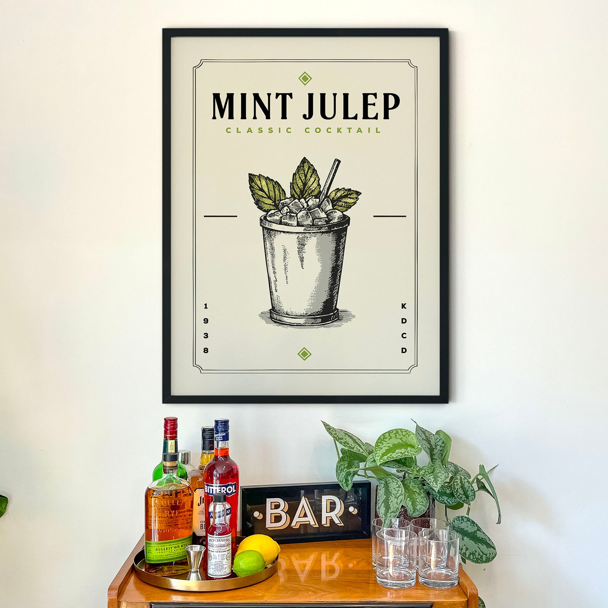 a framed poster of a mint julep cocktail hanging on a wall above a bar cart. The poster includes illustrations of the cocktail and the word "MINT JULEP" prominently displayed.