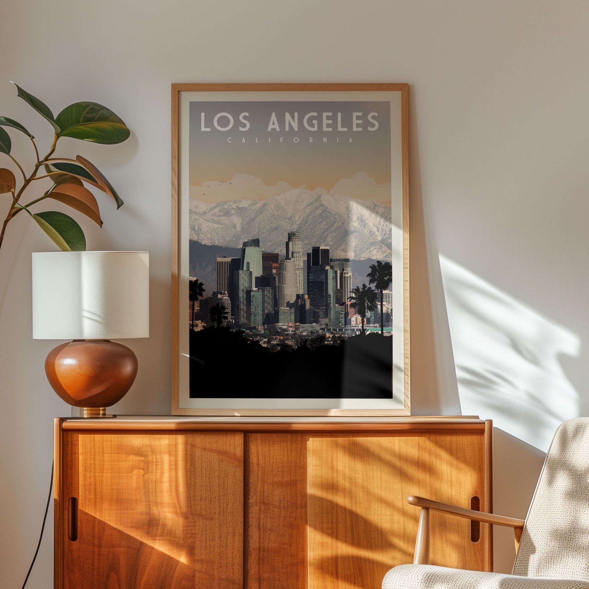 a framed poster of Los Angeles, California, with a cityscape and mountains in the background. The poster is displayed on a wooden cabinet, and there is a lamp and a plant in the foreground.