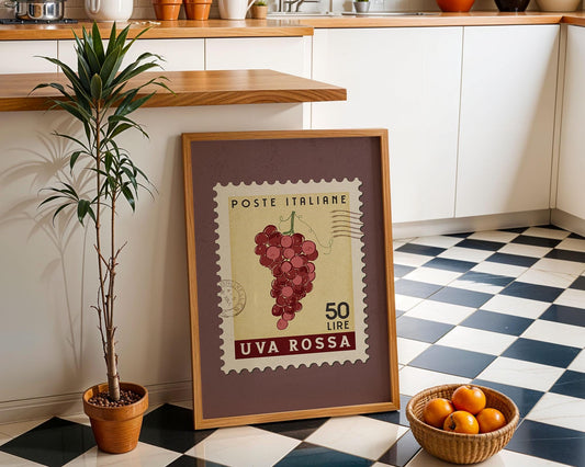 a framed stamp-like artwork depicting a bunch of grapes, placed on a checkered floor next to a potted plant and a basket of oranges.