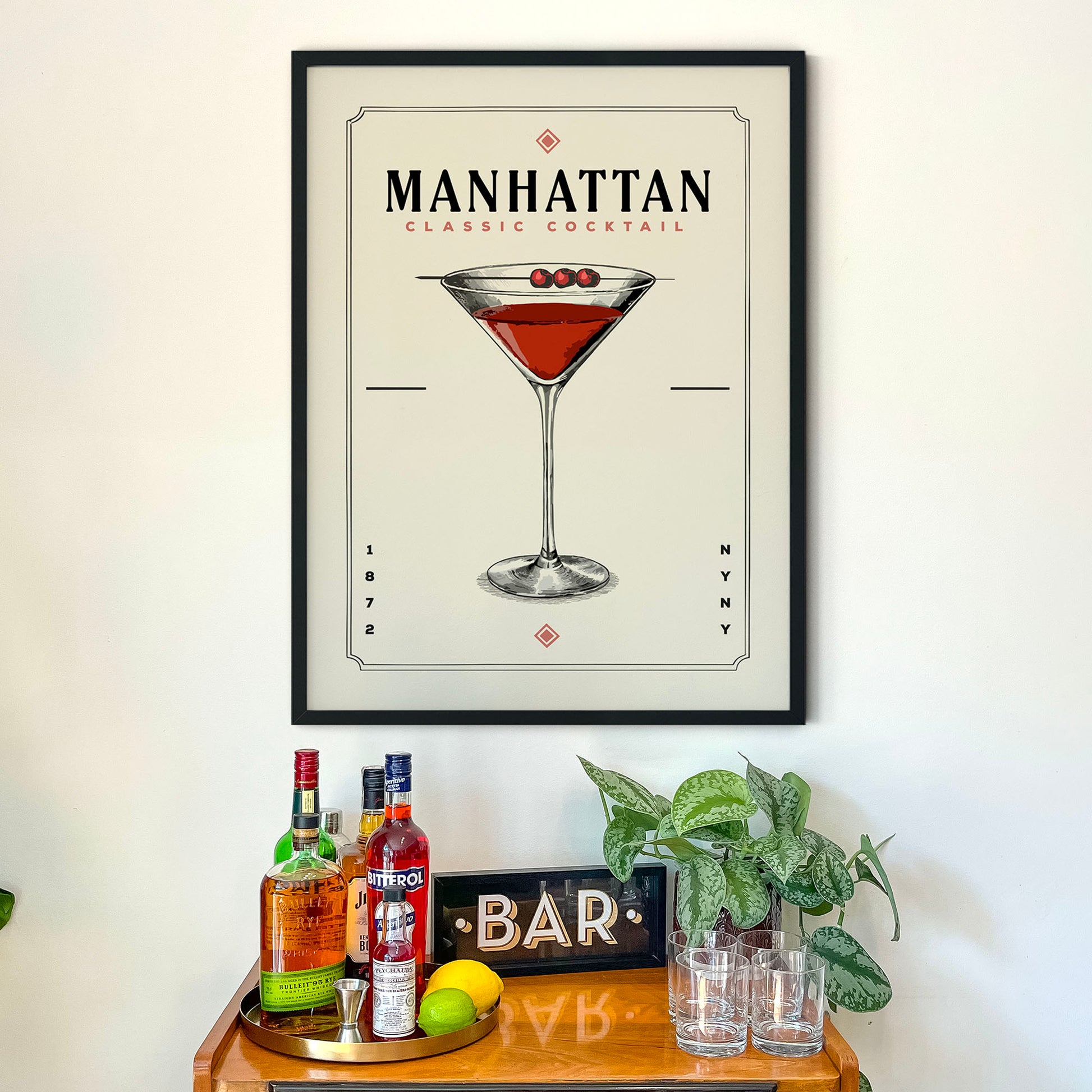 a framed poster of a Manhattan cocktail on a wall above a wooden bar cart with various liquor bottles and glasses.
