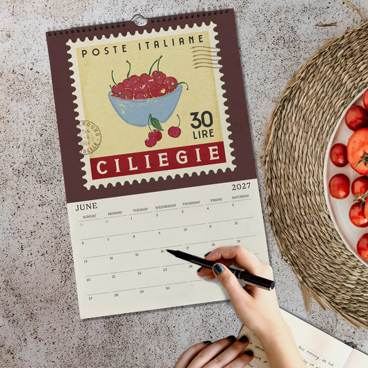 a person's hand holding a pen, writing on a calendar with a vintage-style stamp design. The calendar is titled "Poste Italiane" and displays the date "June" and the location "Ciliegie". The person is also holding a plate of tomatoes, which are visible on the table.