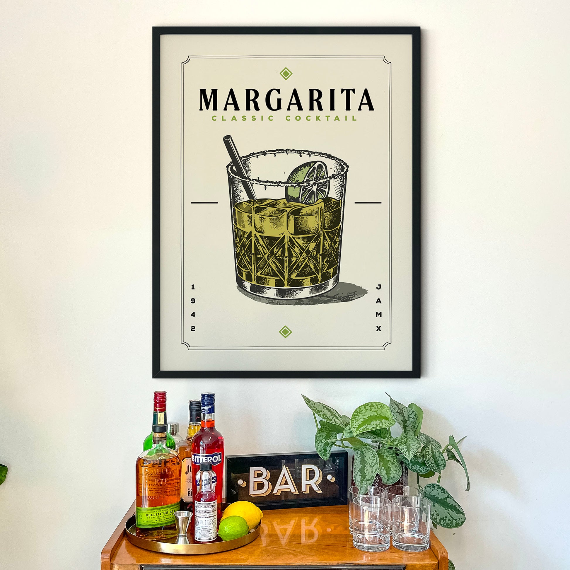 a framed poster of a Margarita cocktail hanging on a wall above a wooden bar cart. The poster displays the Margarita cocktail illustration and includes the text "MARGARITA CLASSIC COCKTAIL" and "SAR" below the illustration. The bar cart is filled with various liquor bottles and has a sign that reads "BAR".