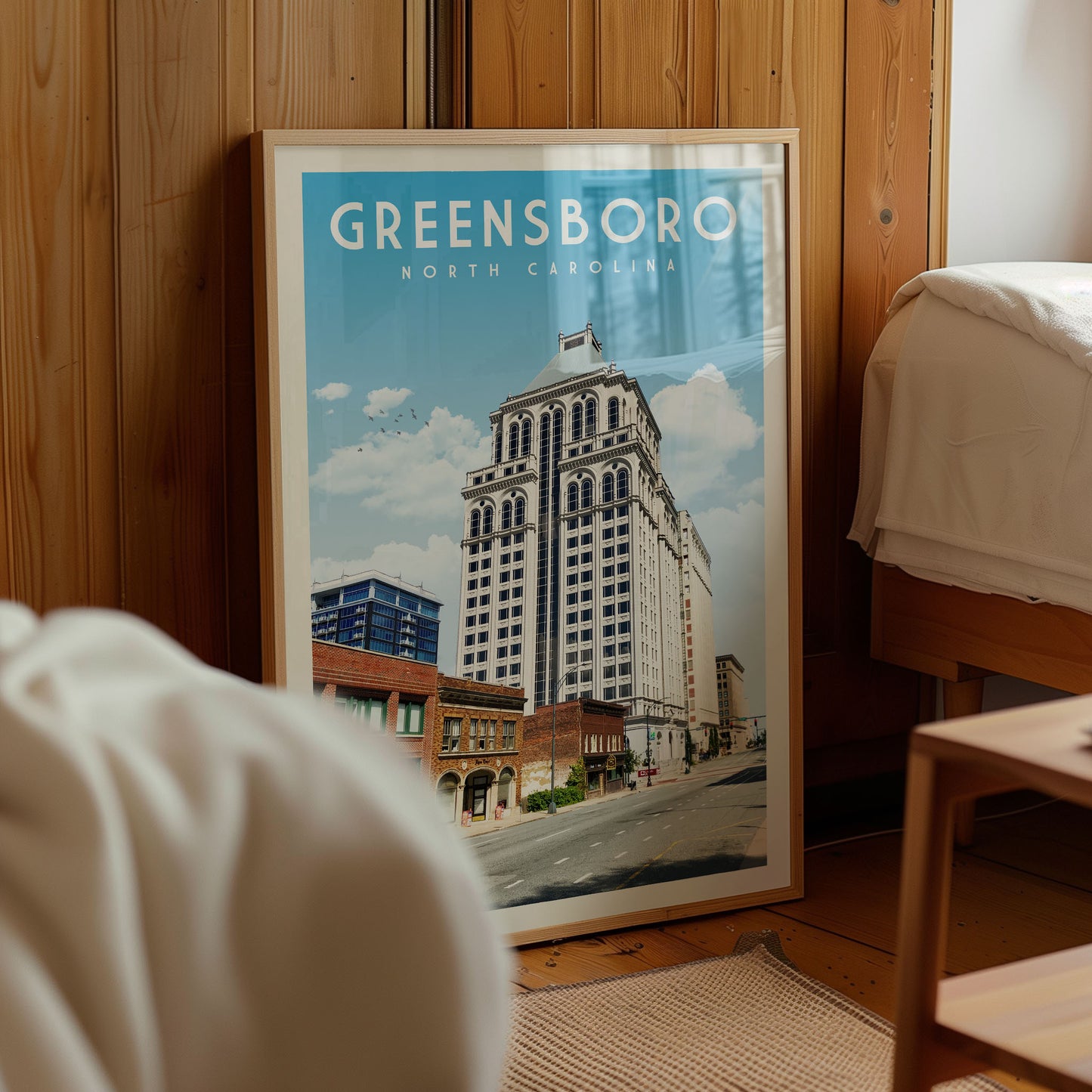 A framed poster of Greensboro, North Carolina, is displayed in a room with a wooden wall and a bed visible in the background.