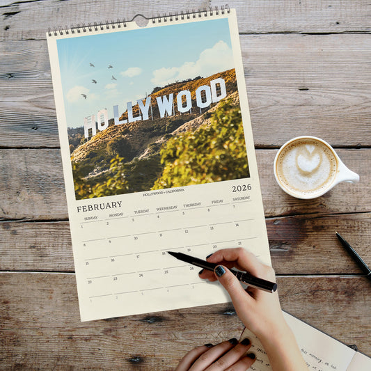 A person's hand is writing in a notebook while holding a pen, with a calendar for February displayed on a wooden surface next to a cup of coffee.