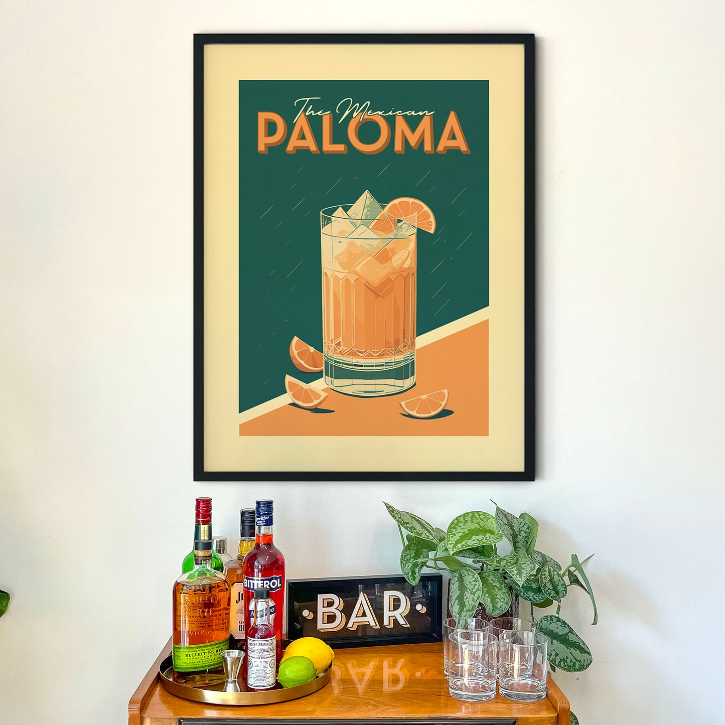 a framed poster of a cocktail glass with a drink, a wooden bar cart with various bottles of alcohol, and a potted plant.