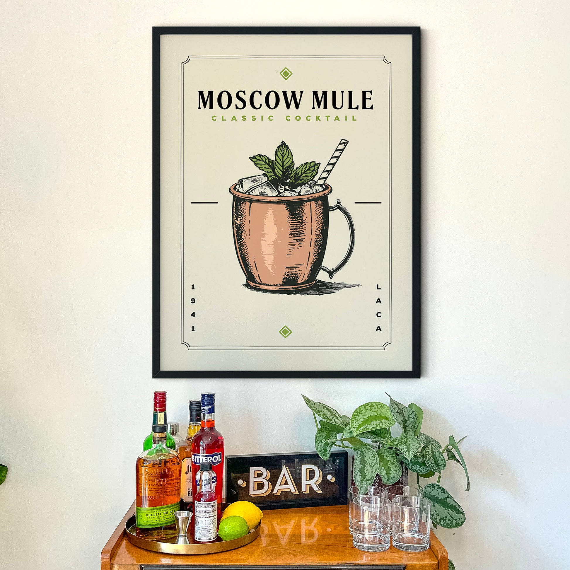 a framed poster of a Moscow Mule cocktail hanging on a wall above a wooden bar cart. The poster displays the Moscow Mule cocktail with a copper mug and a mint sprig. The bar cart has various bottles of alcohol and glasses, creating a cozy and inviting atmosphere.
