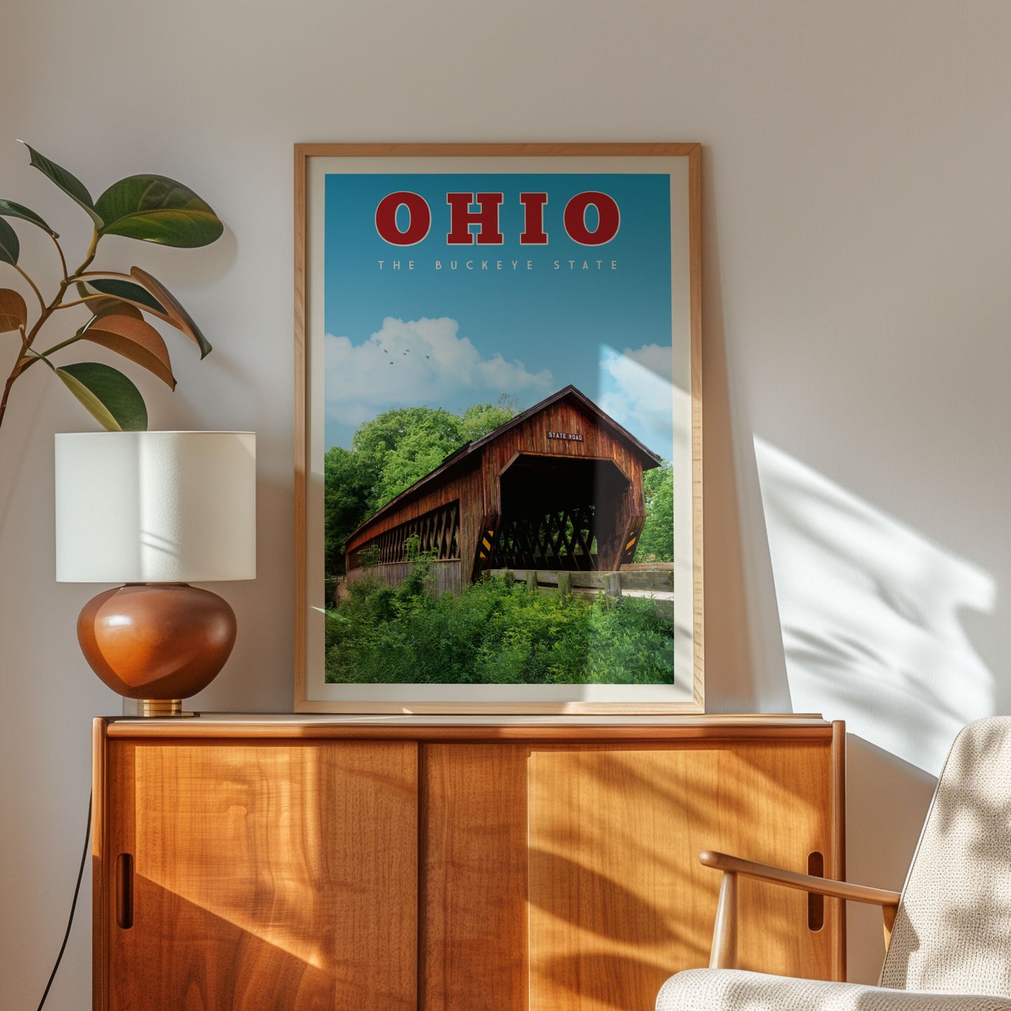 a framed poster of a covered bridge in Ohio, with a wooden cabinet below it and a lamp on a side table.
