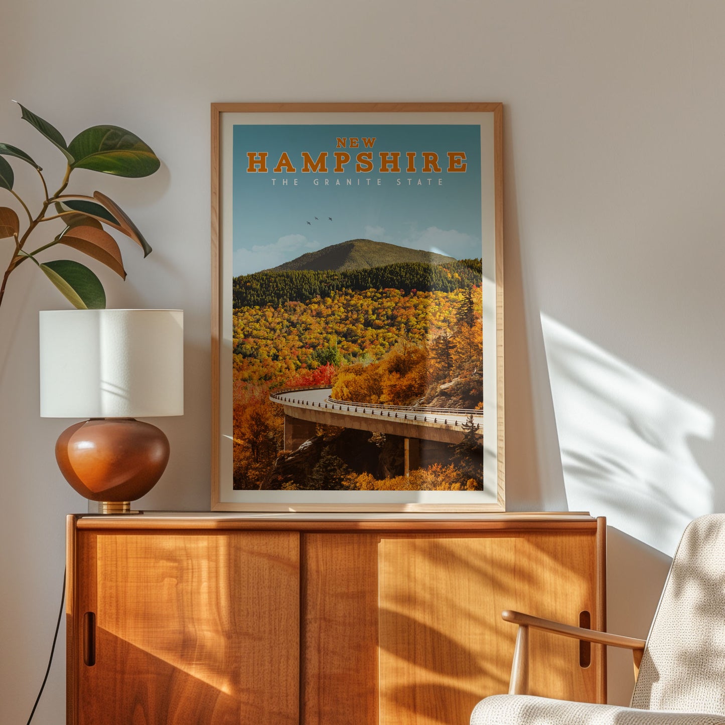 a framed poster of a scenic autumn landscape with a bridge, hanging on a wall next to a wooden cabinet and a lamp.