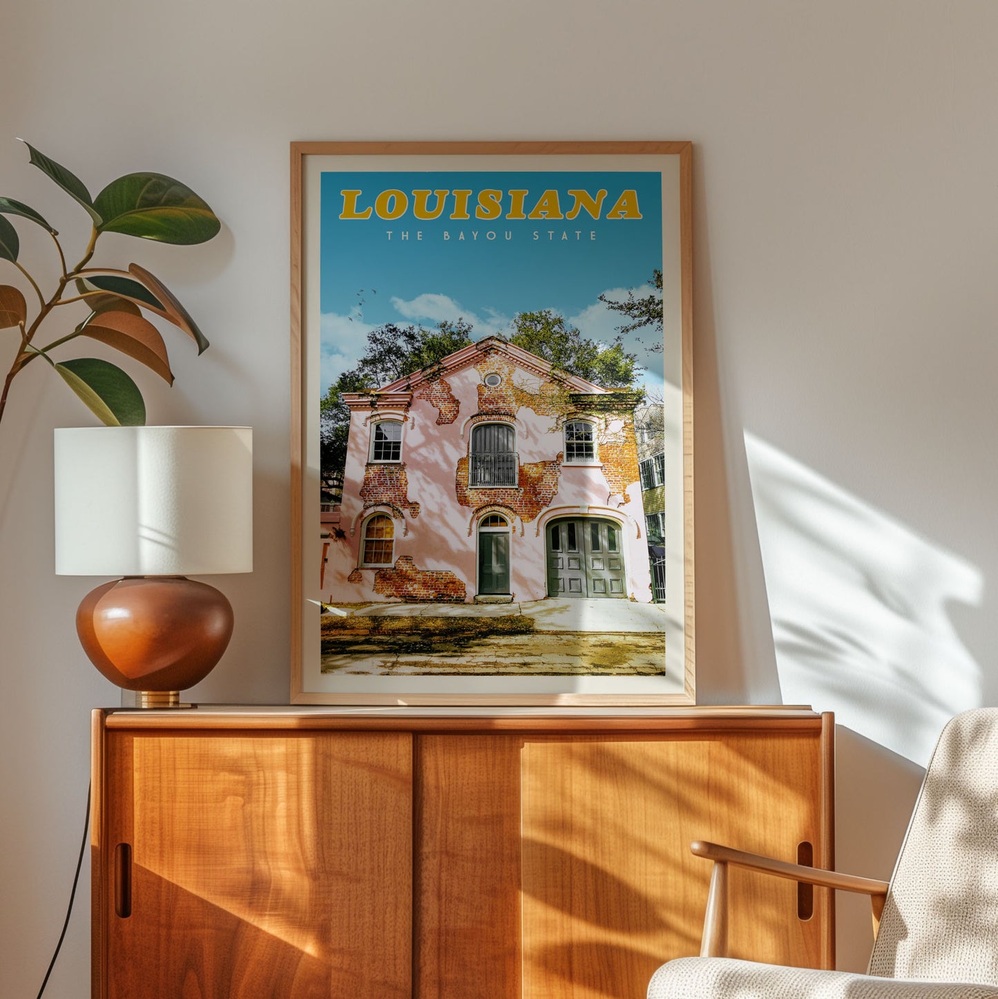 a framed poster of a building in Louisiana, with a wooden cabinet below it and a lamp on a side table.