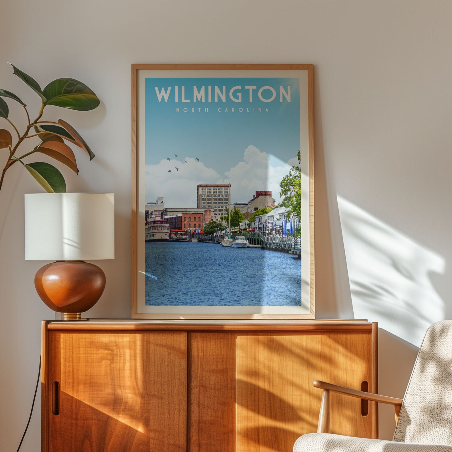 A framed poster of a cityscape with a river in the background, hanging on a wall next to a wooden cabinet and a lamp.