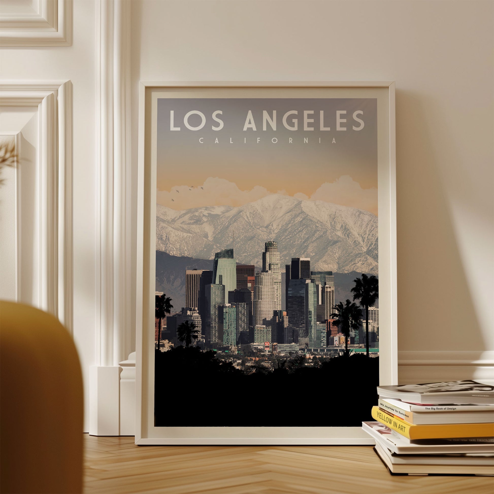 A framed poster of the Los Angeles skyline with mountains in the background, placed on a wooden floor next to a stack of books.
