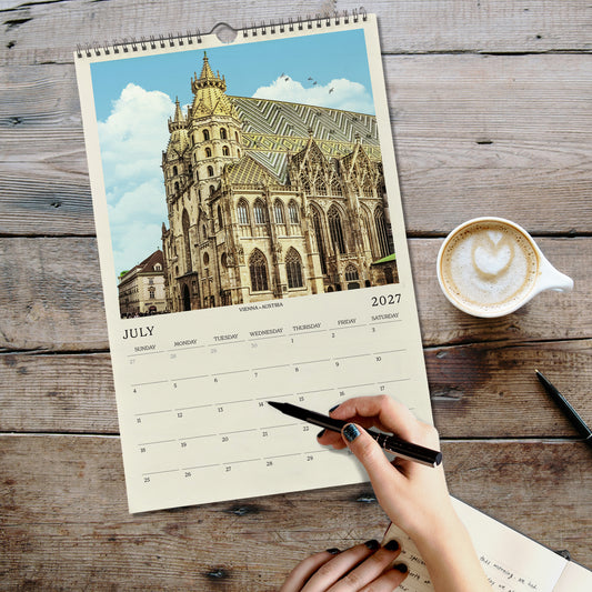A person's hand is writing in a notebook while holding a pen, with a coffee cup and a spiral-bound calendar for July displayed on a wooden surface.