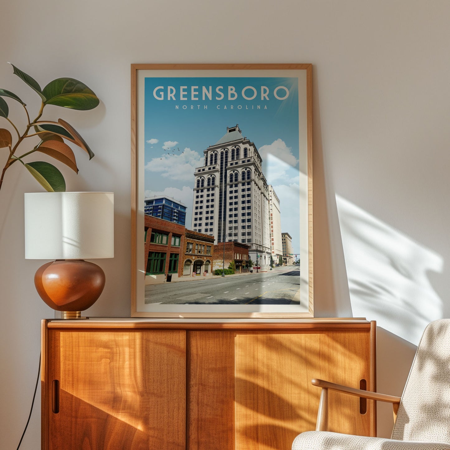 a framed poster of Greensboro, North Carolina, with a cityscape of tall buildings and a street in the background. The poster is displayed on a wooden cabinet, and there is a lamp and a plant in the foreground.