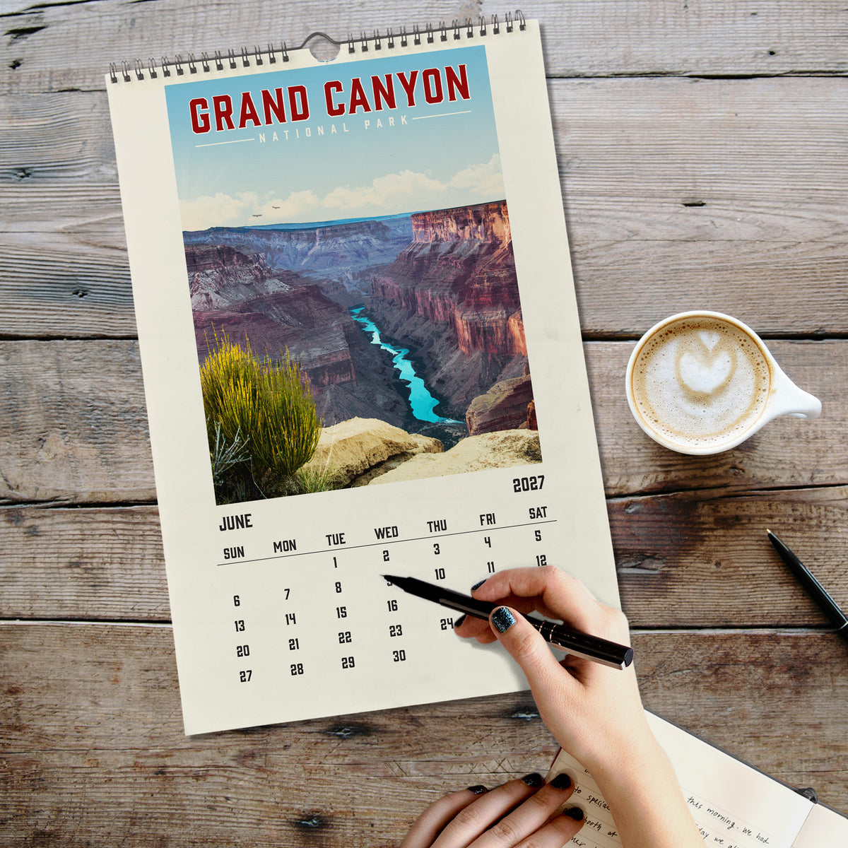 A person's hand is writing in a notebook on a wooden surface, with a calendar for Grand Canyon National Park displayed nearby.
