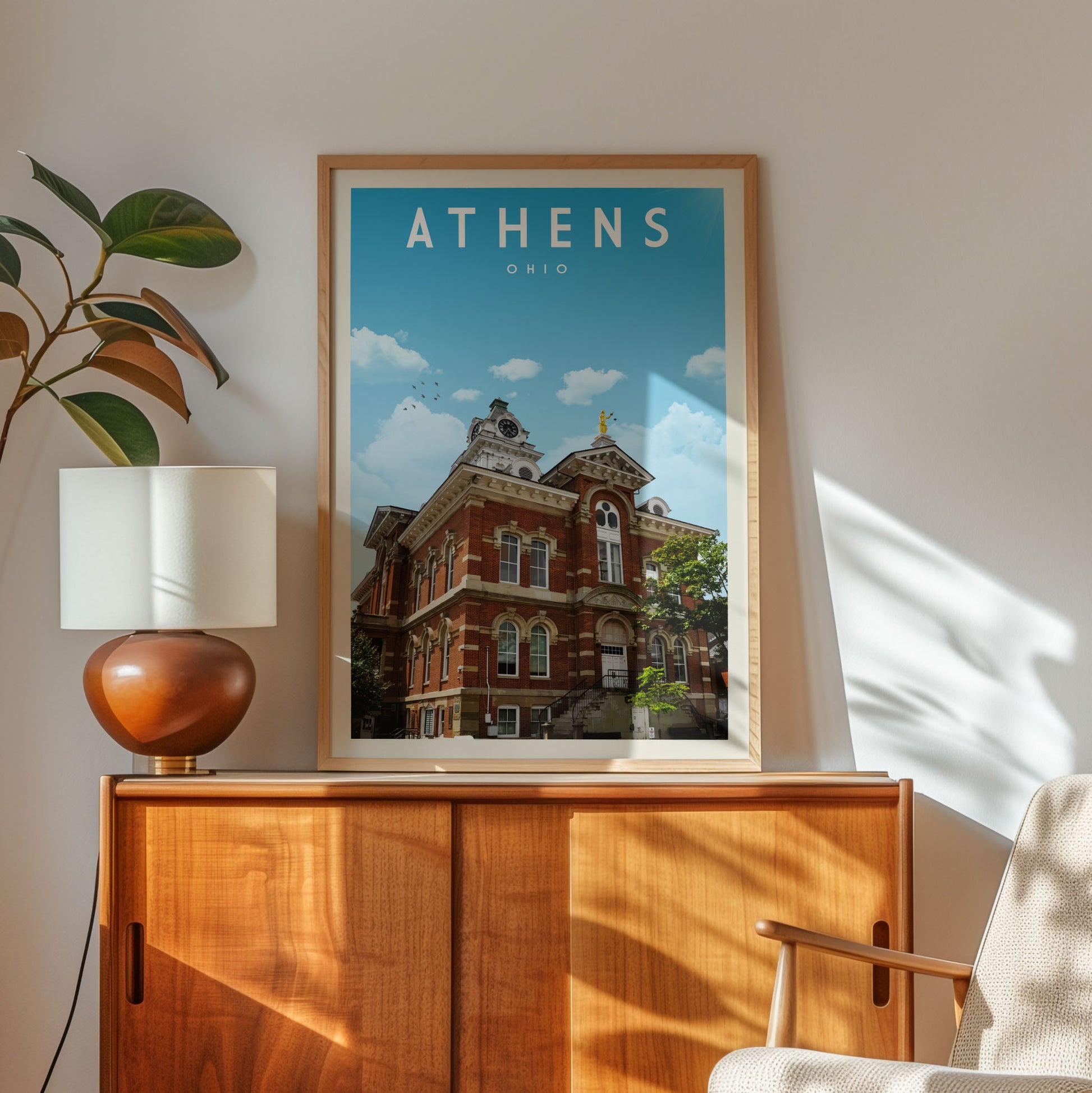a framed poster of a building with the word "ATHENS" written on it, hanging on a wall next to a wooden cabinet and a lamp.
