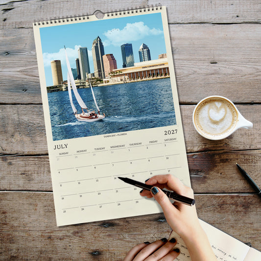 A person's hand is writing in a notebook while holding a pen, with a spiral-bound calendar featuring a cityscape and a sailboat on the cover.