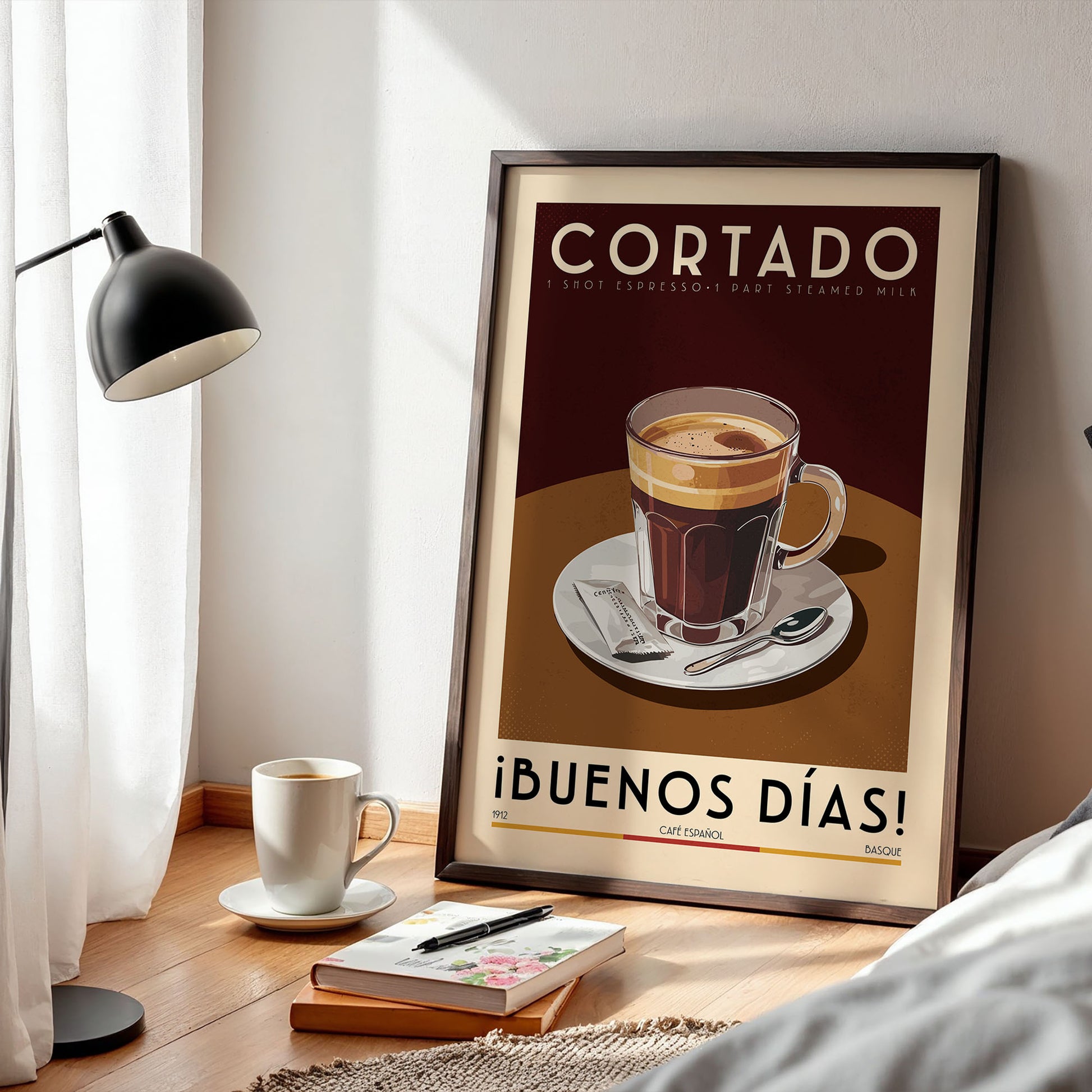 a framed poster of a cup of coffee with the text "Cortado" and "iBUENOS DÍAS!" on it, along with a cup of coffee and a book on a wooden table.