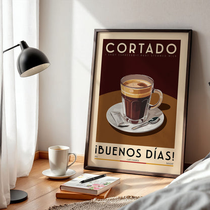 a framed poster of a cup of coffee with the text "Cortado" and "iBUENOS DÍAS!" on it, along with a cup of coffee and a book on a wooden table.