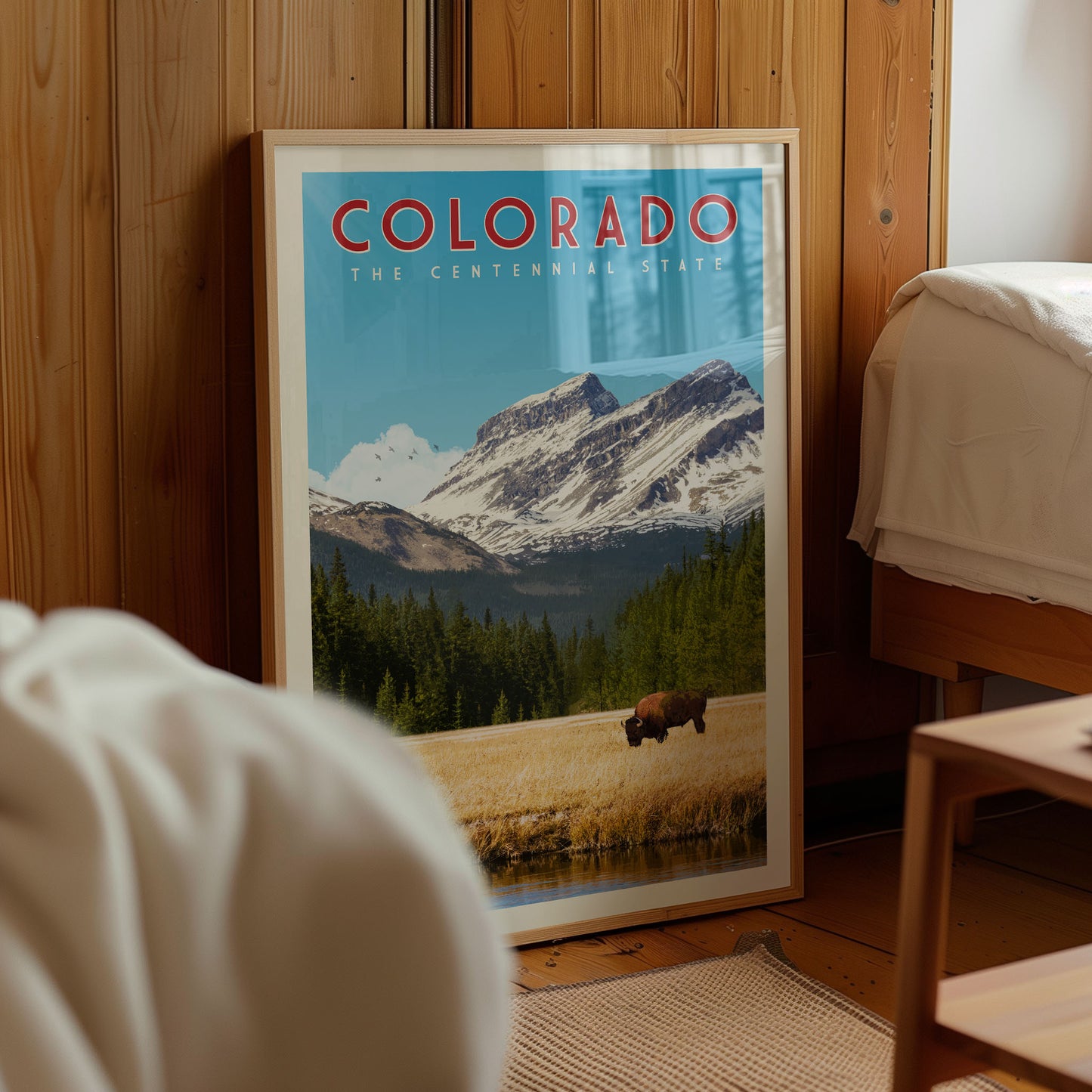 A framed poster of a scenic mountain landscape with a bison grazing in the foreground, displayed in a room with wooden walls.