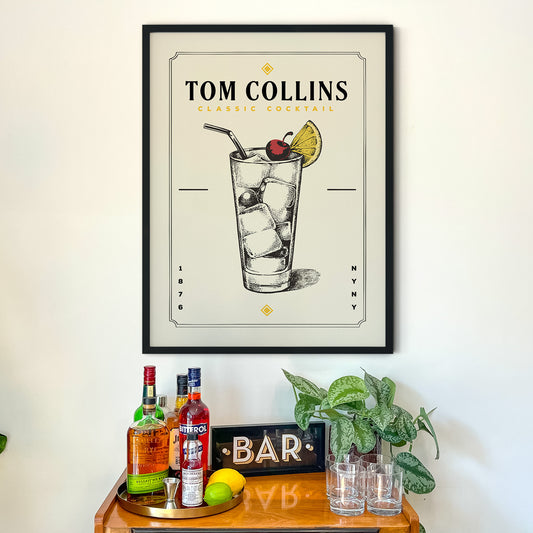 a framed poster of a cocktail on a wall above a bar counter with various bottles of alcohol and glasses.