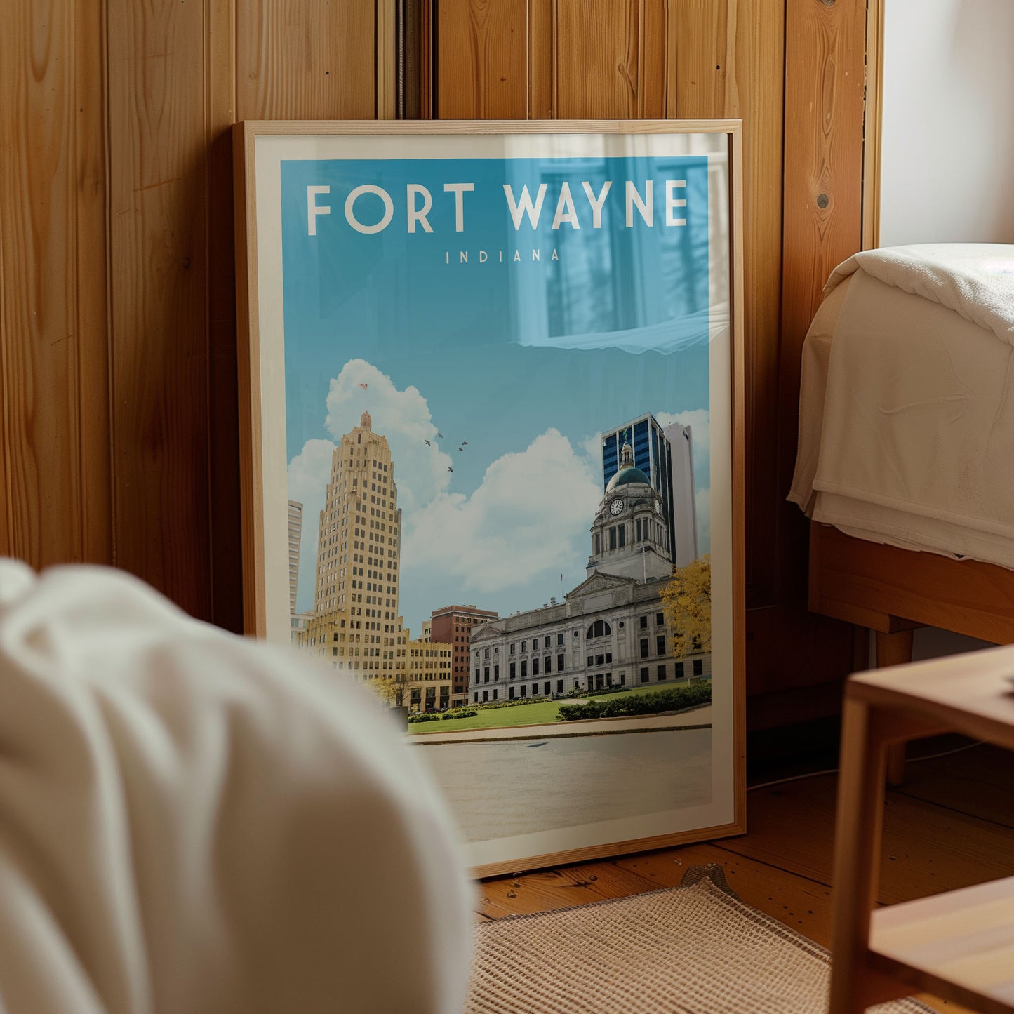 A framed poster of Fort Wayne, Indiana, is displayed in a room with a wooden wall and a bed visible in the background.
