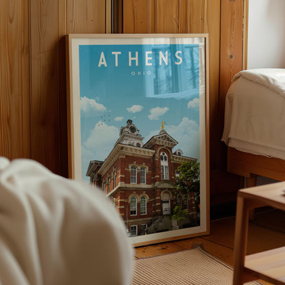 A framed poster of a historic building in Athens, Ohio, displayed in a room with a wooden wall and a bed visible in the background.