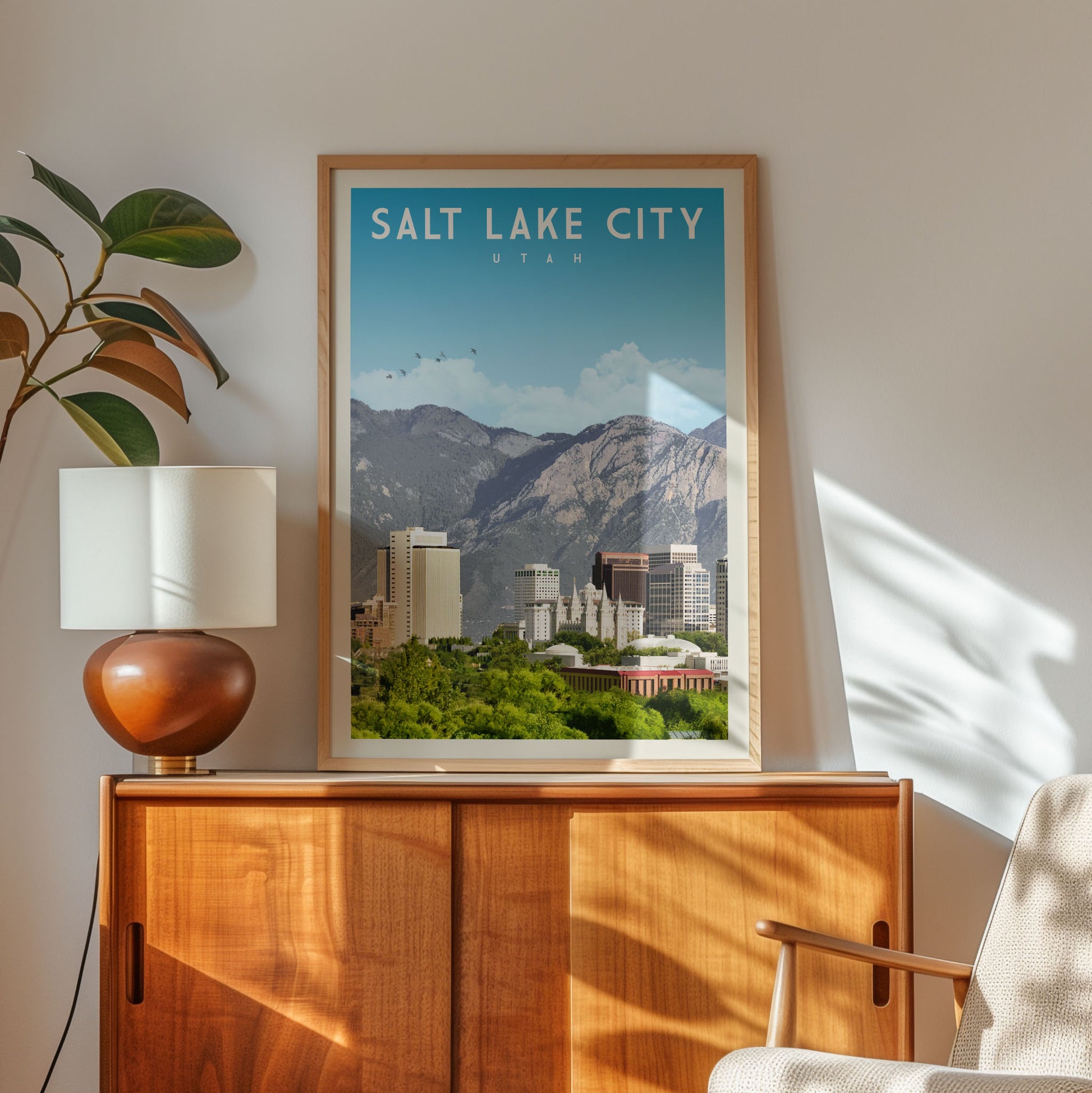 a framed poster of Salt Lake City, Utah, with a mountain landscape in the background and a cityscape in the foreground. The poster is displayed on a wooden cabinet, and there is a lamp and a plant in the foreground.
