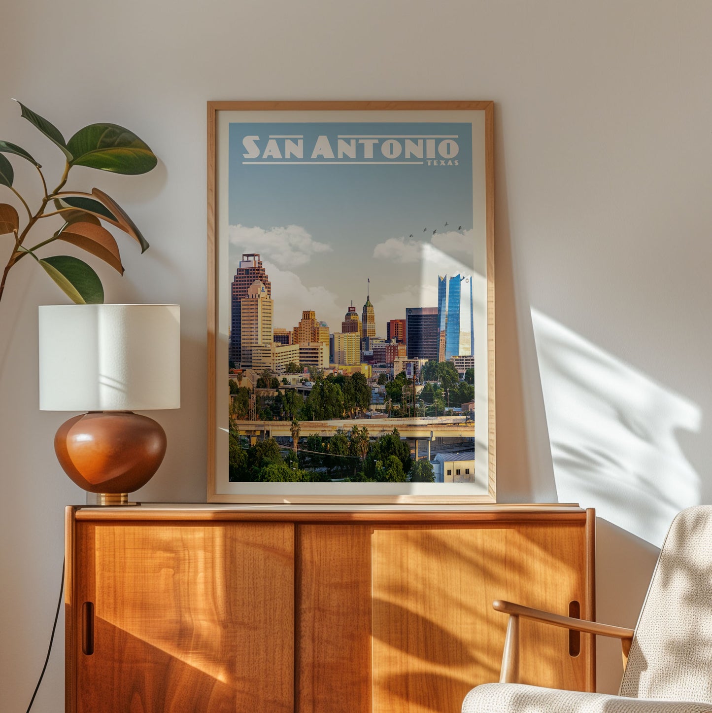 a framed poster of the San Antonio skyline, with a wooden cabinet and a lamp in the foreground.