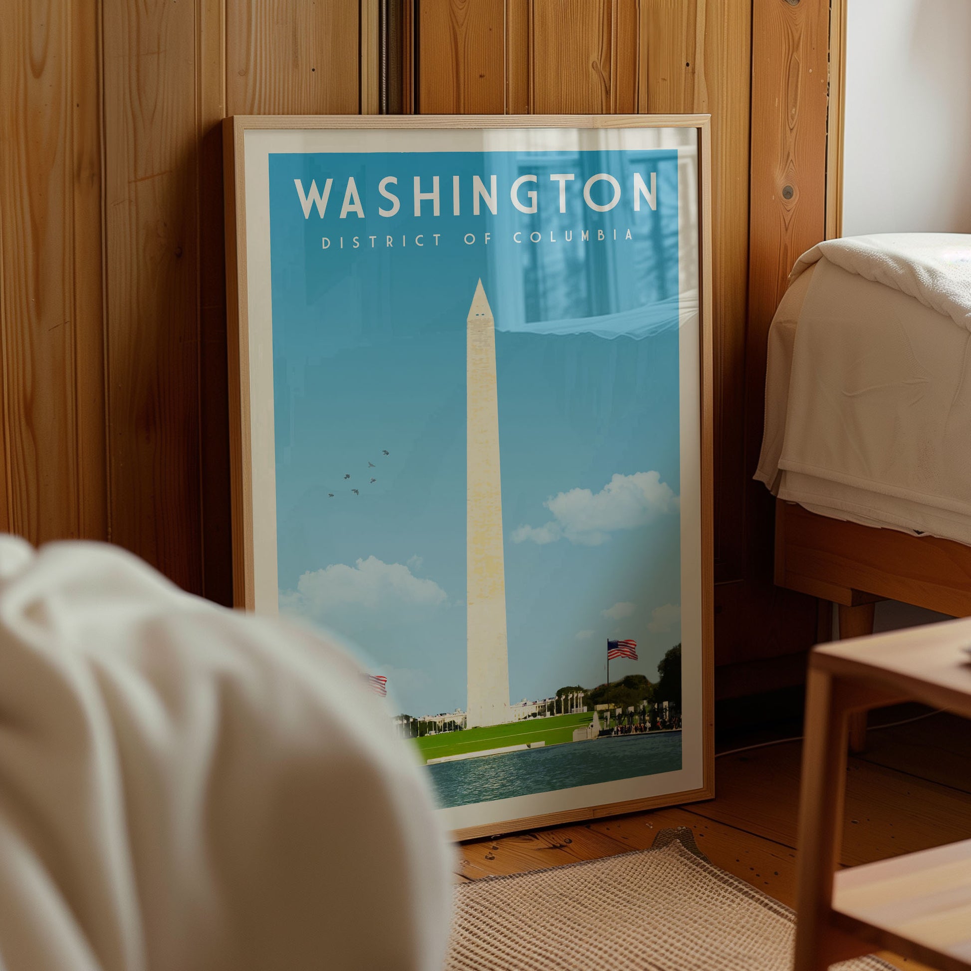 A framed poster of the Washington Monument is displayed in a room with a wooden wall and a bed visible in the background.