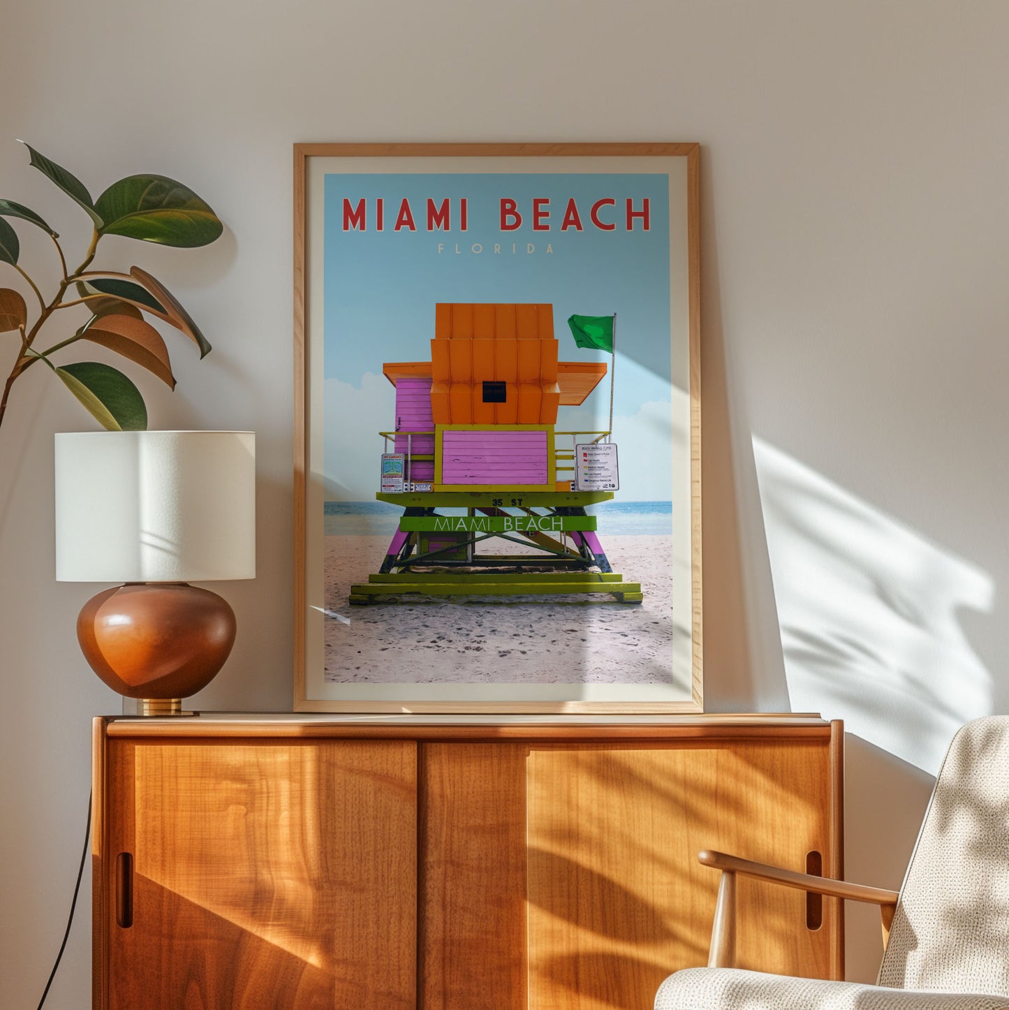 a framed poster of a colorful beach house, with the text "MIAMI BEACH" prominently displayed. The poster is hanging on a wall, and there is a lamp and a plant in the foreground.