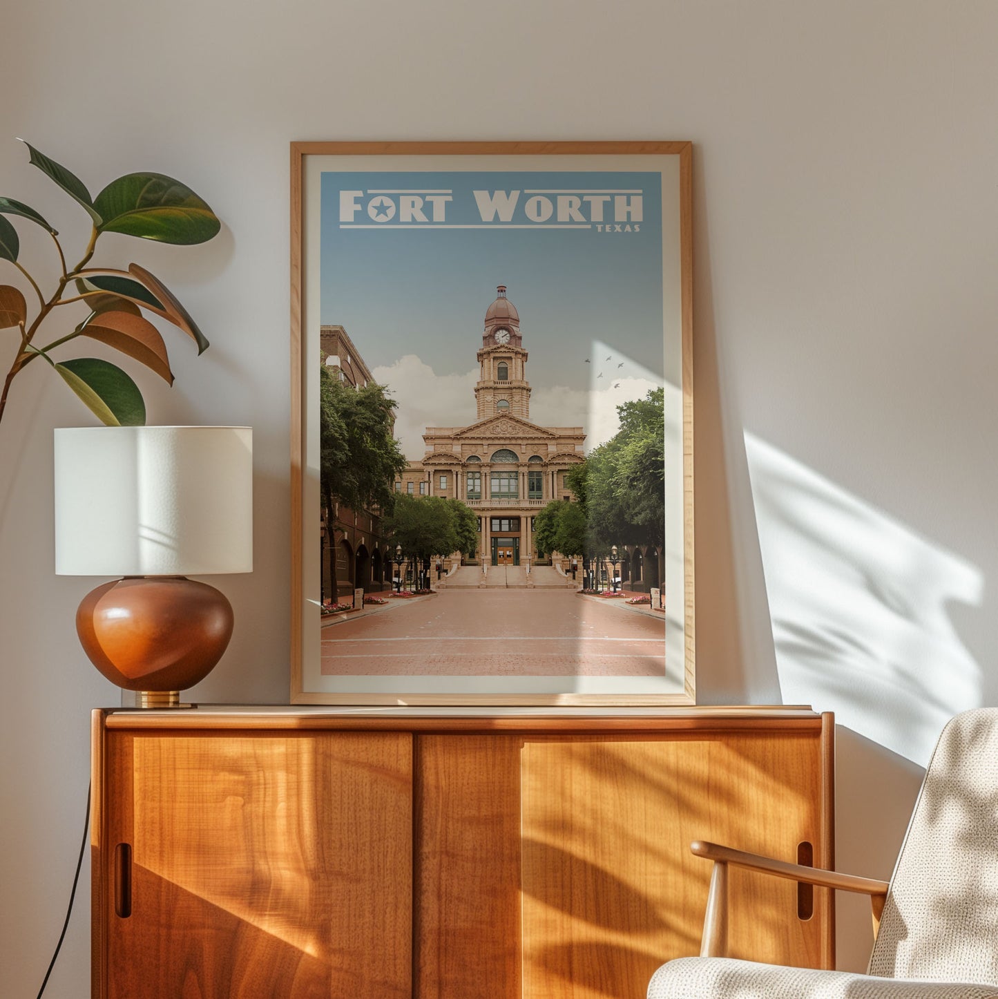 a framed poster of Fort Worth, Texas, with a clock tower and a street scene in the background. The poster is displayed on a wooden cabinet, and there is a lamp and a plant in the foreground.