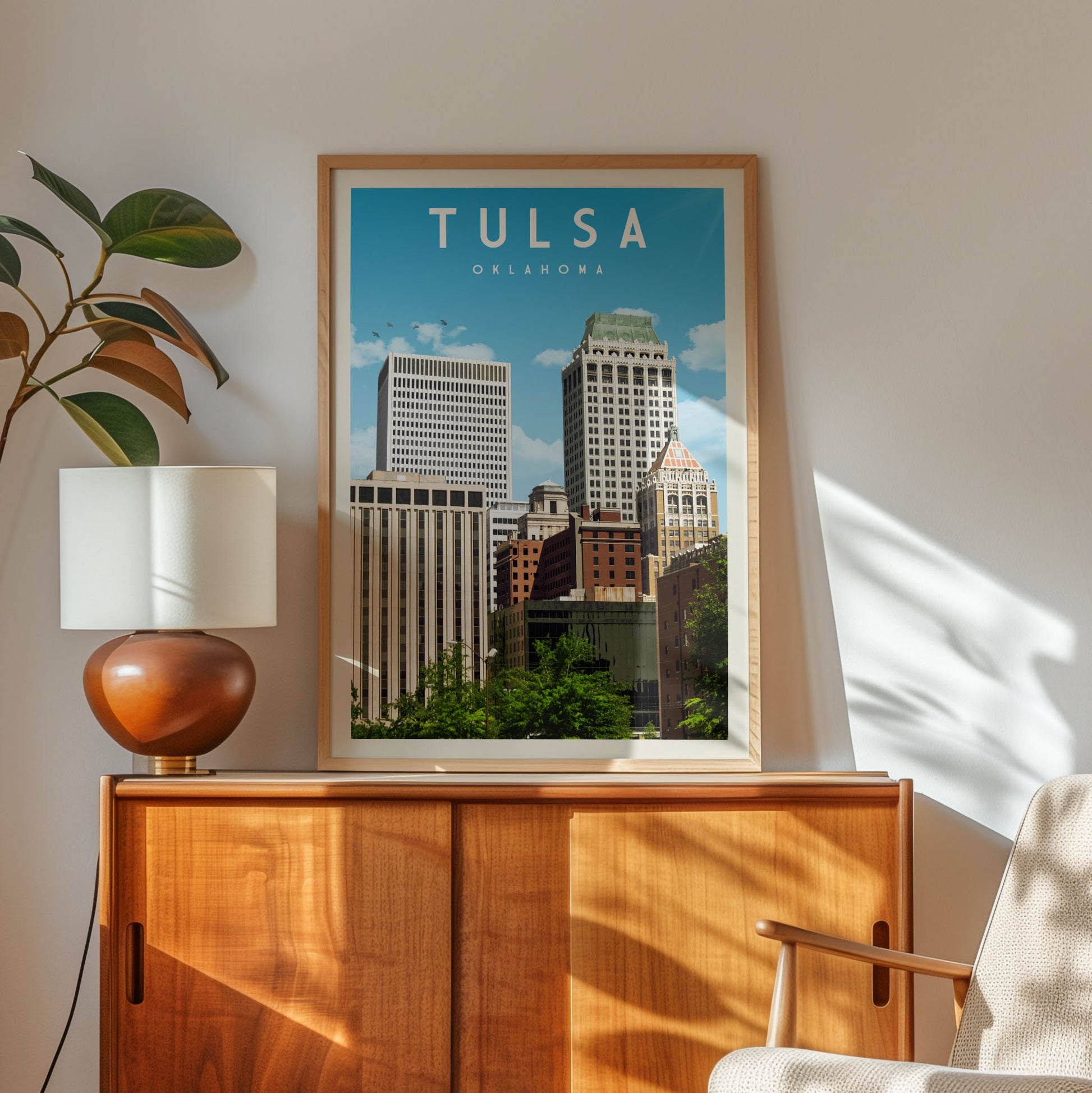 a framed poster of a cityscape of Oklahoma, with two tall buildings prominently displayed. The poster is hung on a wooden cabinet, and there is a lamp and a plant in the foreground.