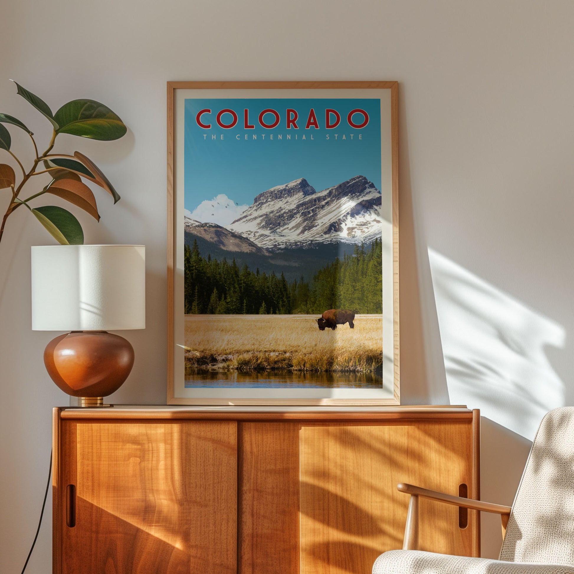 a framed poster of a scenic mountain landscape with a cow grazing in the foreground, placed on a wooden cabinet in a room with a lamp and a plant.