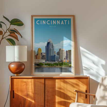 a framed poster of the Cincinnati skyline, with a wooden cabinet and a lamp in the foreground.