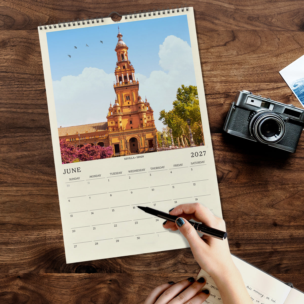 A person's hand is holding a pen and writing on a calendar, with a camera and other items on a wooden surface.