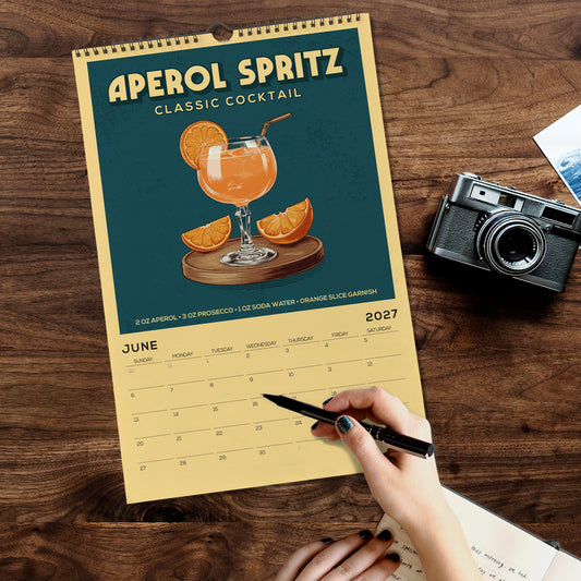 A person is writing on a calendar with a pen, surrounded by a camera, a notebook, and a glass of orange juice on a wooden table.