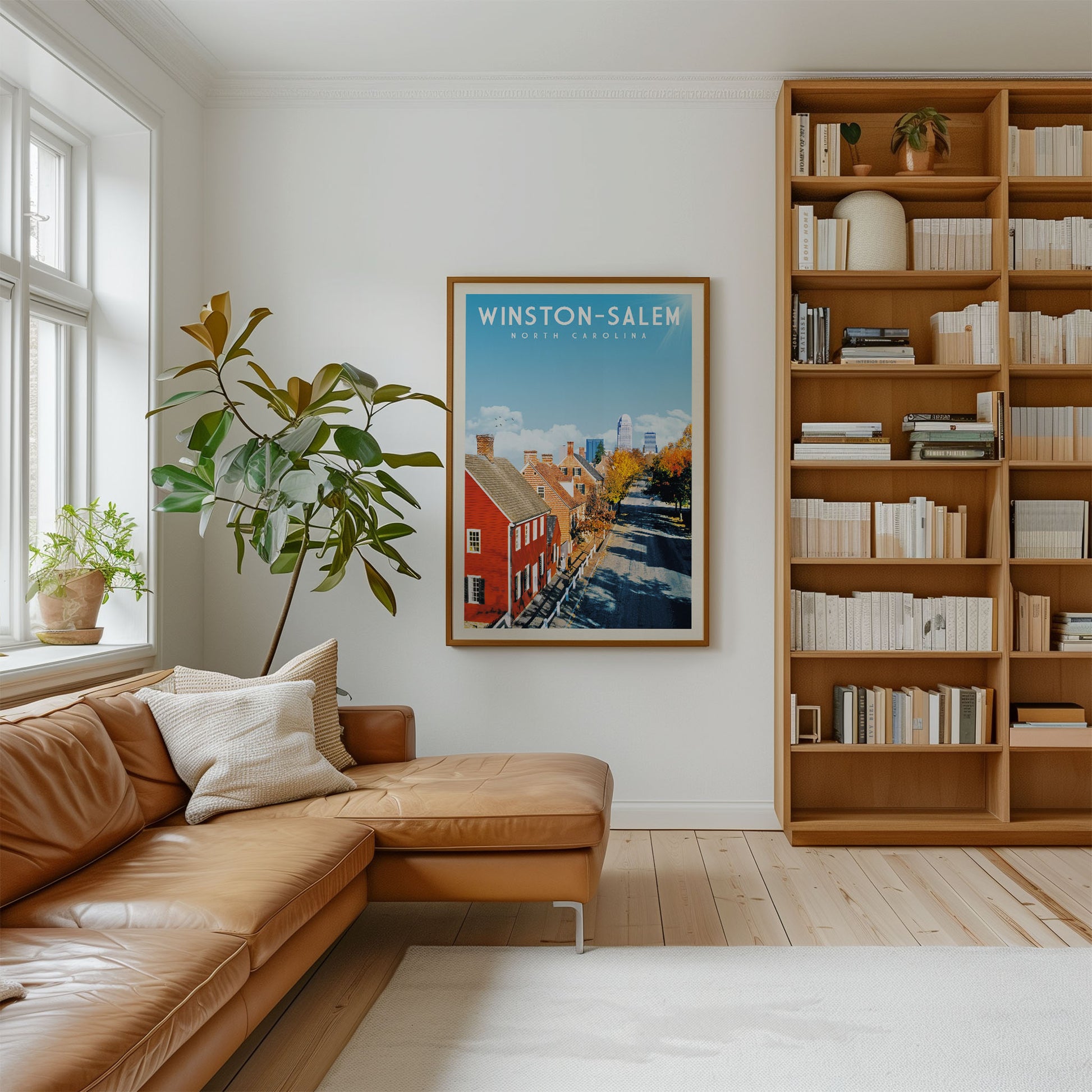 a cozy living room with a large window, a brown leather sofa, a wooden bookshelf filled with books, and a framed poster of Winston-Salem, North Carolina.