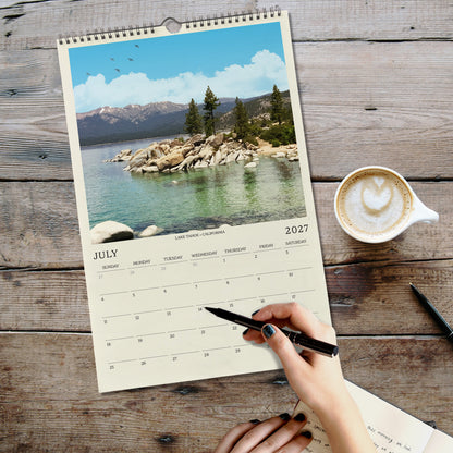 A person's hand is writing in a notebook while holding a pen, with a coffee cup and a scenic landscape visible on a wooden table.