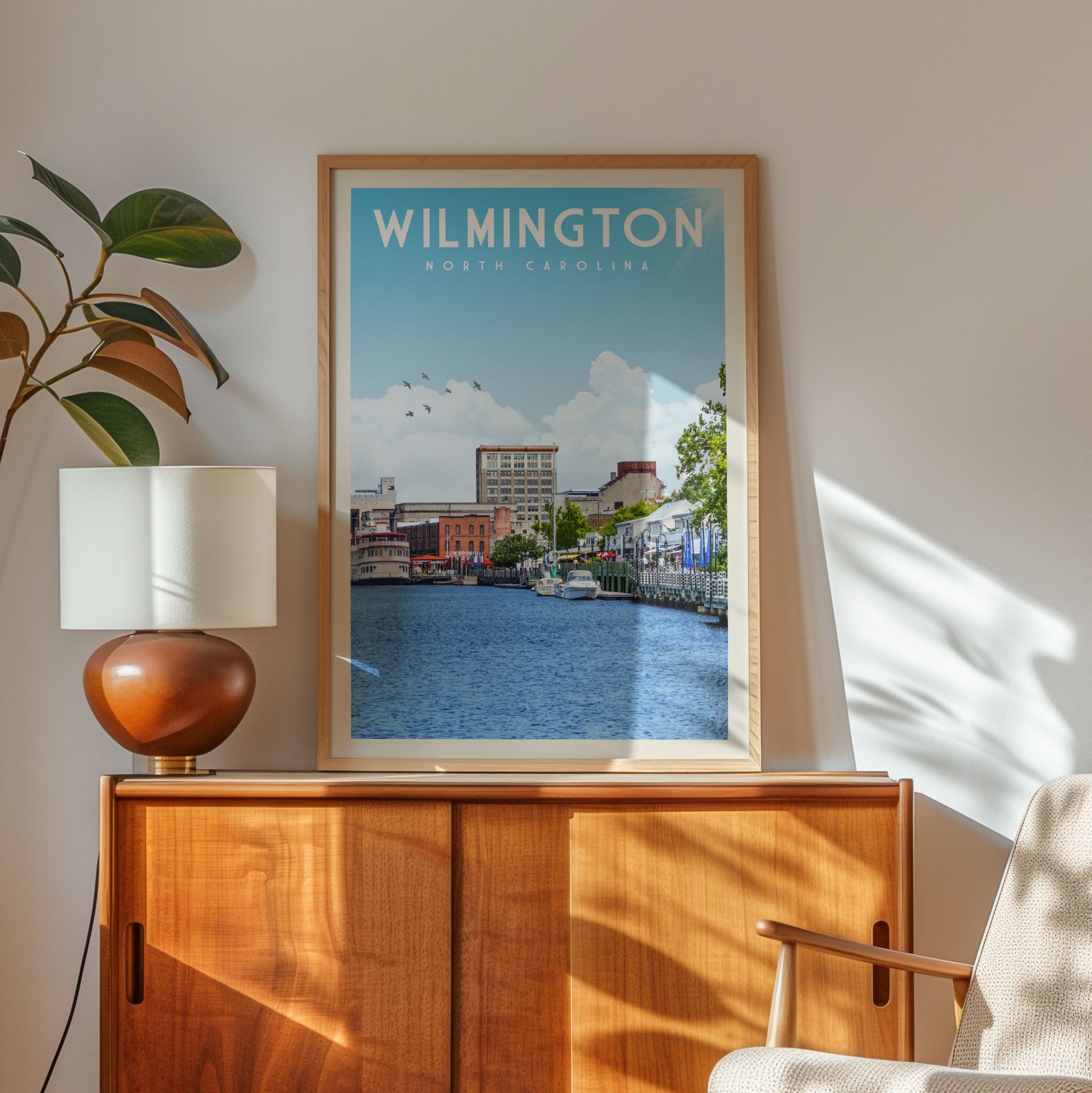 A framed poster of a cityscape with a river in the background, hanging on a wall next to a wooden cabinet and a lamp.