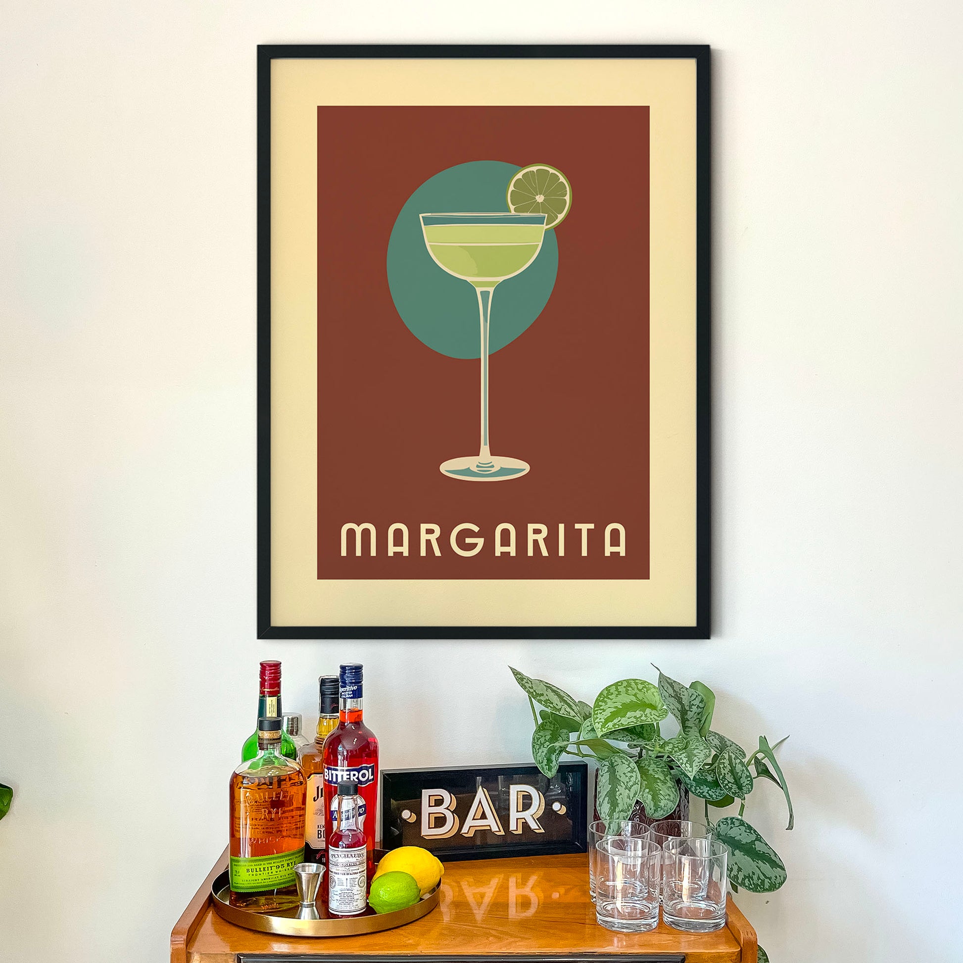 a framed poster of a margarita glass above a wooden bar cart with various bottles of alcohol and a potted plant.