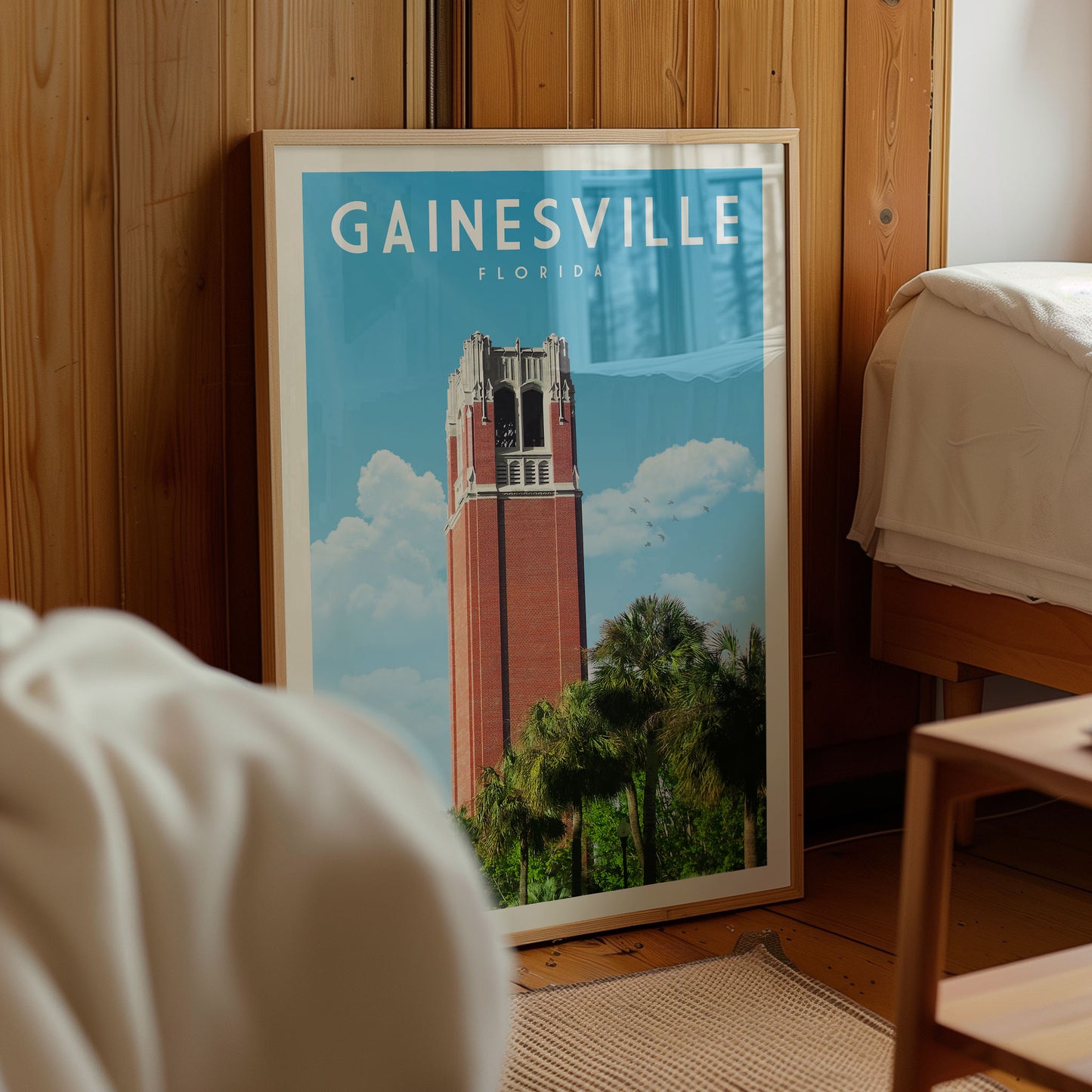A framed poster of the Gainesville, Florida skyline, featuring the iconic Bell Tower, is displayed in a room with a wooden wall and a bed visible in the background.