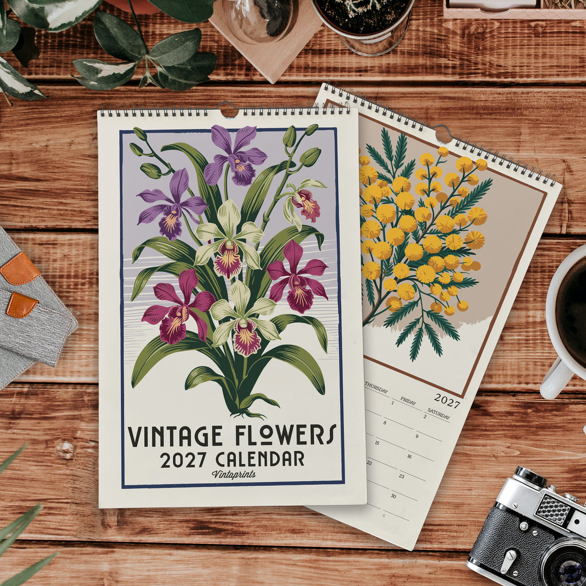 a vintage-style wall calendar with a floral design, placed on a wooden surface alongside other items such as a camera, a mug, and some plants.